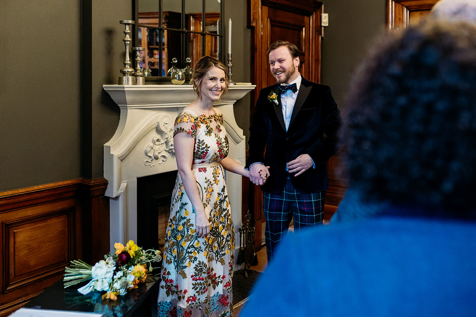 a bride and groom are standing side by side holding hands and smiling. her bouquet is sitting on a table beside them.