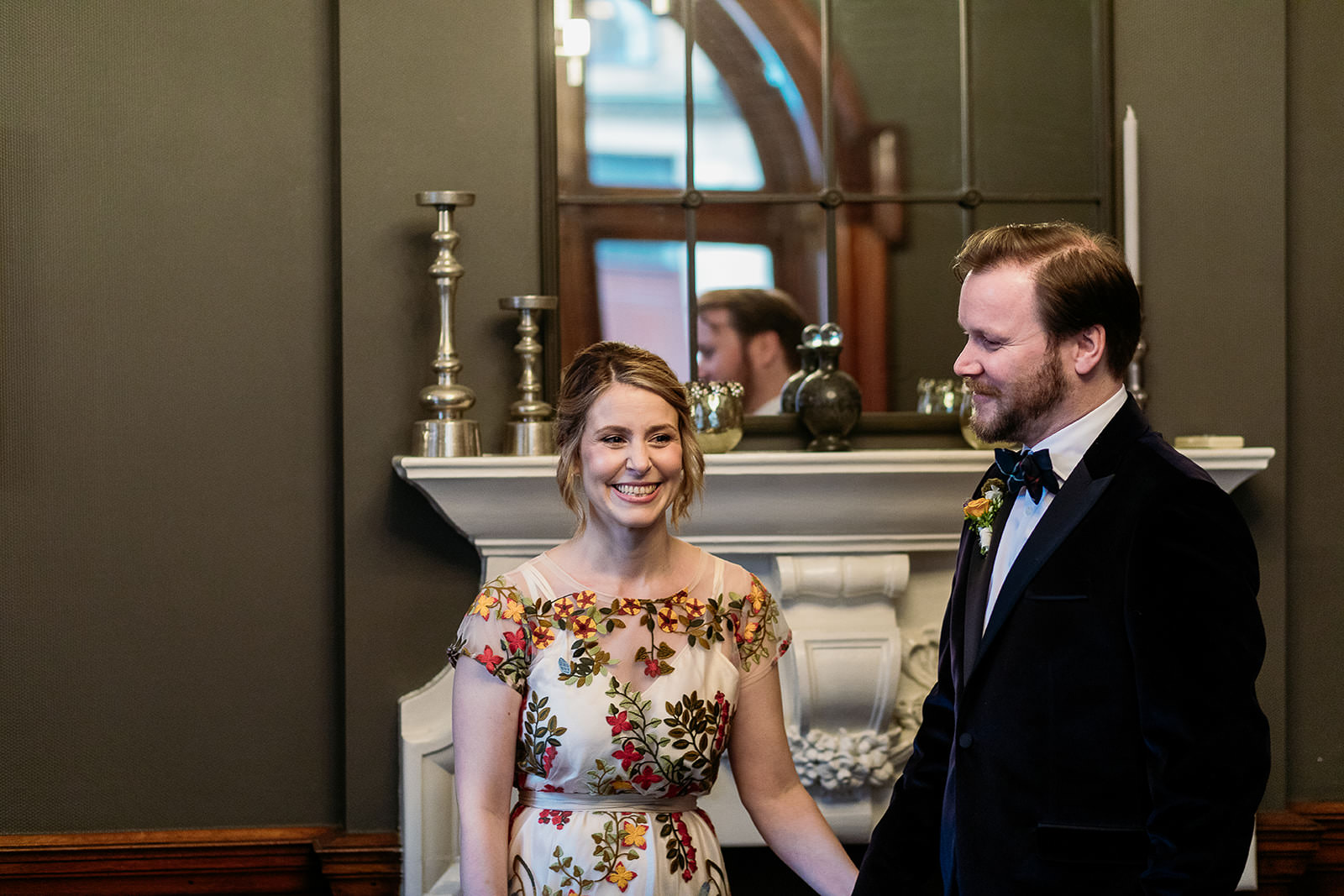 a man and woman are standing together and smiling. behind them is a large white fireplace with silver candlesticks on the mantle. there is a mirror above the fireplace.