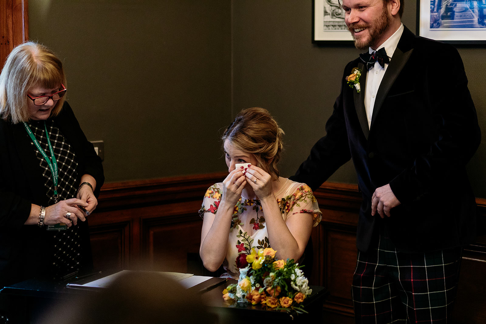 a bride is sitting at a table and she is dabbing under her eye with a tissue. her husband is standing behind his wife leaning on her chair.