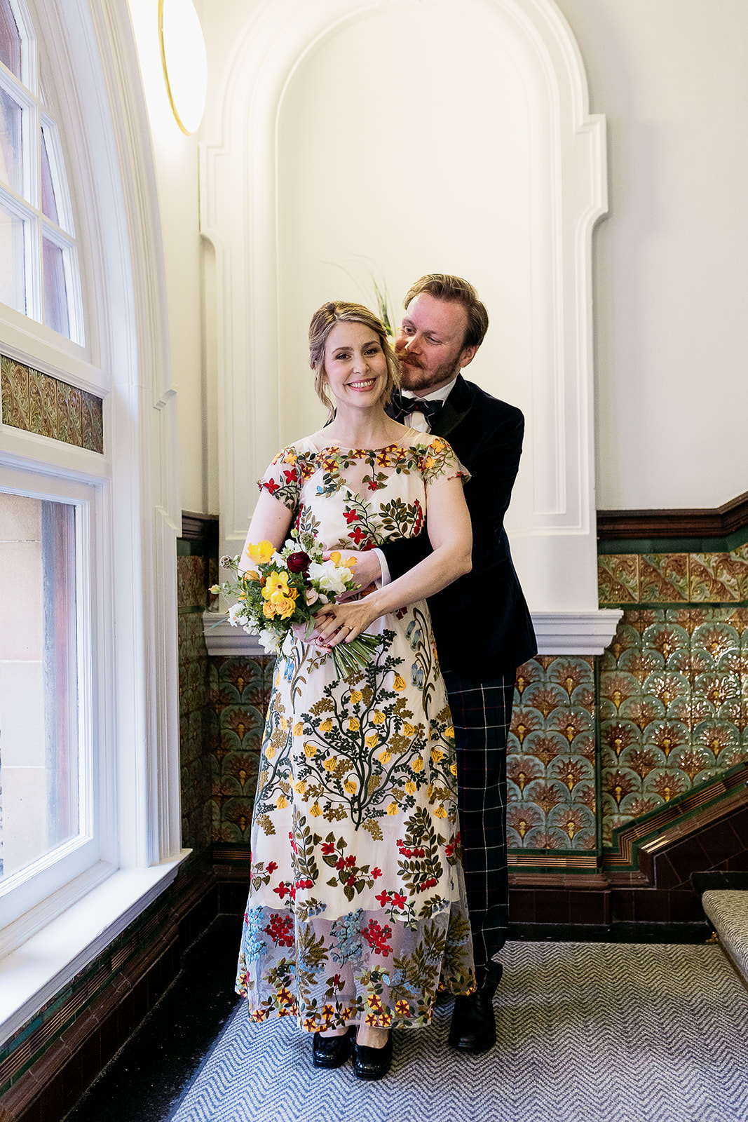 a groom is standing behind his wife, with his arms around her waist. his wife is smiling and he is looking at her.