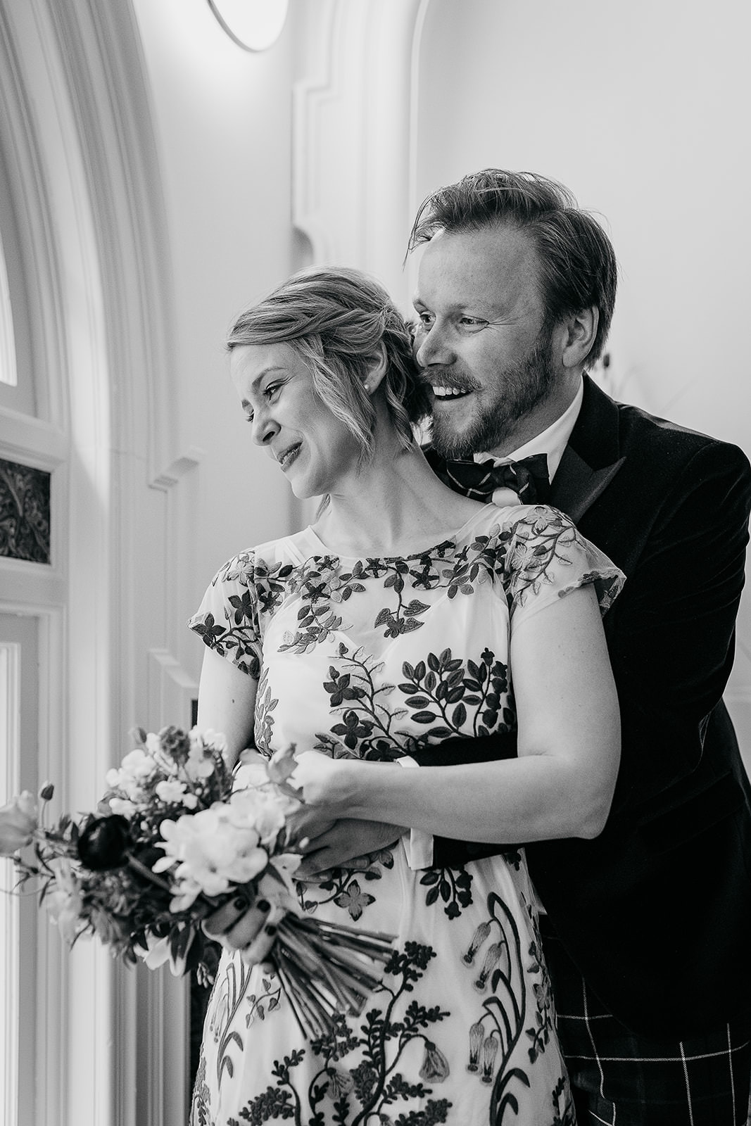 a groom is standing behind his wife and they are looking out of the window. they are both smiling.
