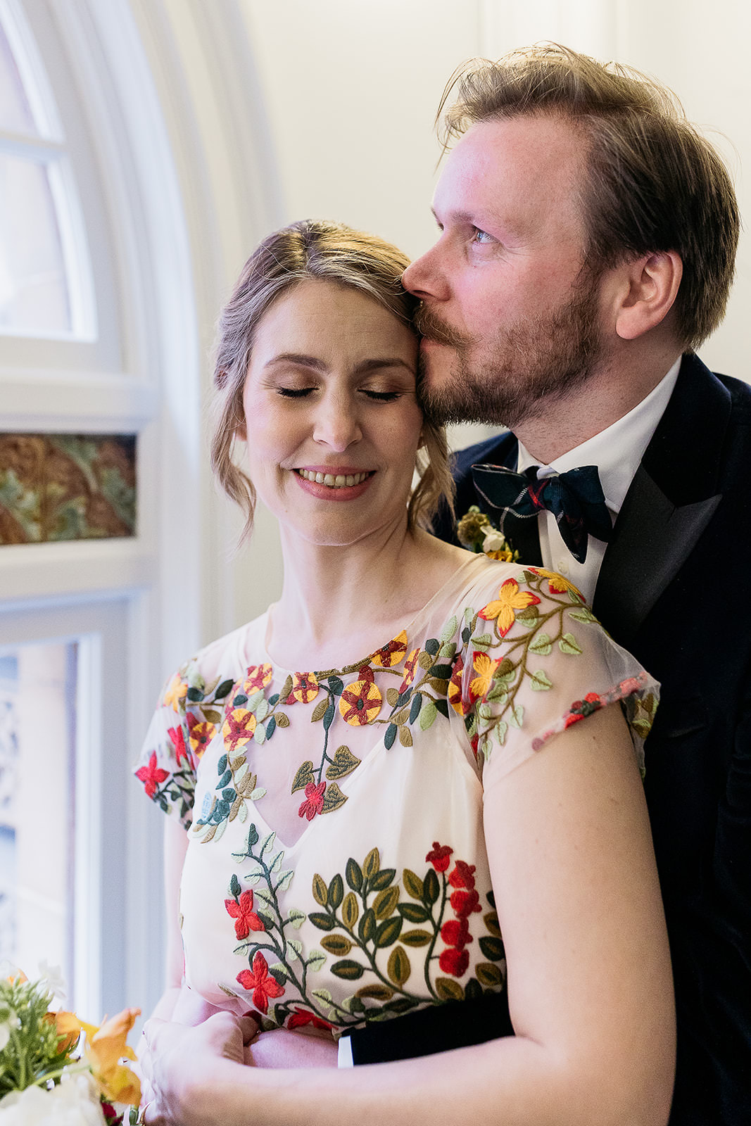 a groom is standing behind his wife and he is looking out of the window while resting his face on the temple of his wife's face. his wife is smiling and has her eyes closed.