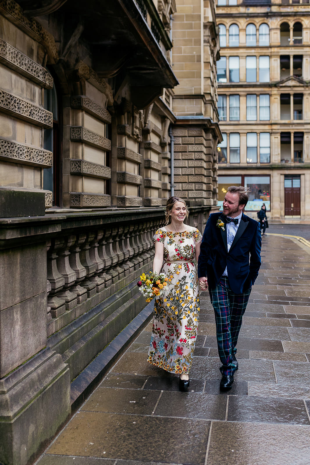 a bride and groom are walking down a street in Glasgow city centre. They are holding hands and smiling. the bride's dress is embroidered with colourful flowers and the groom is wearing tartan trews and a blue velvet jacket.