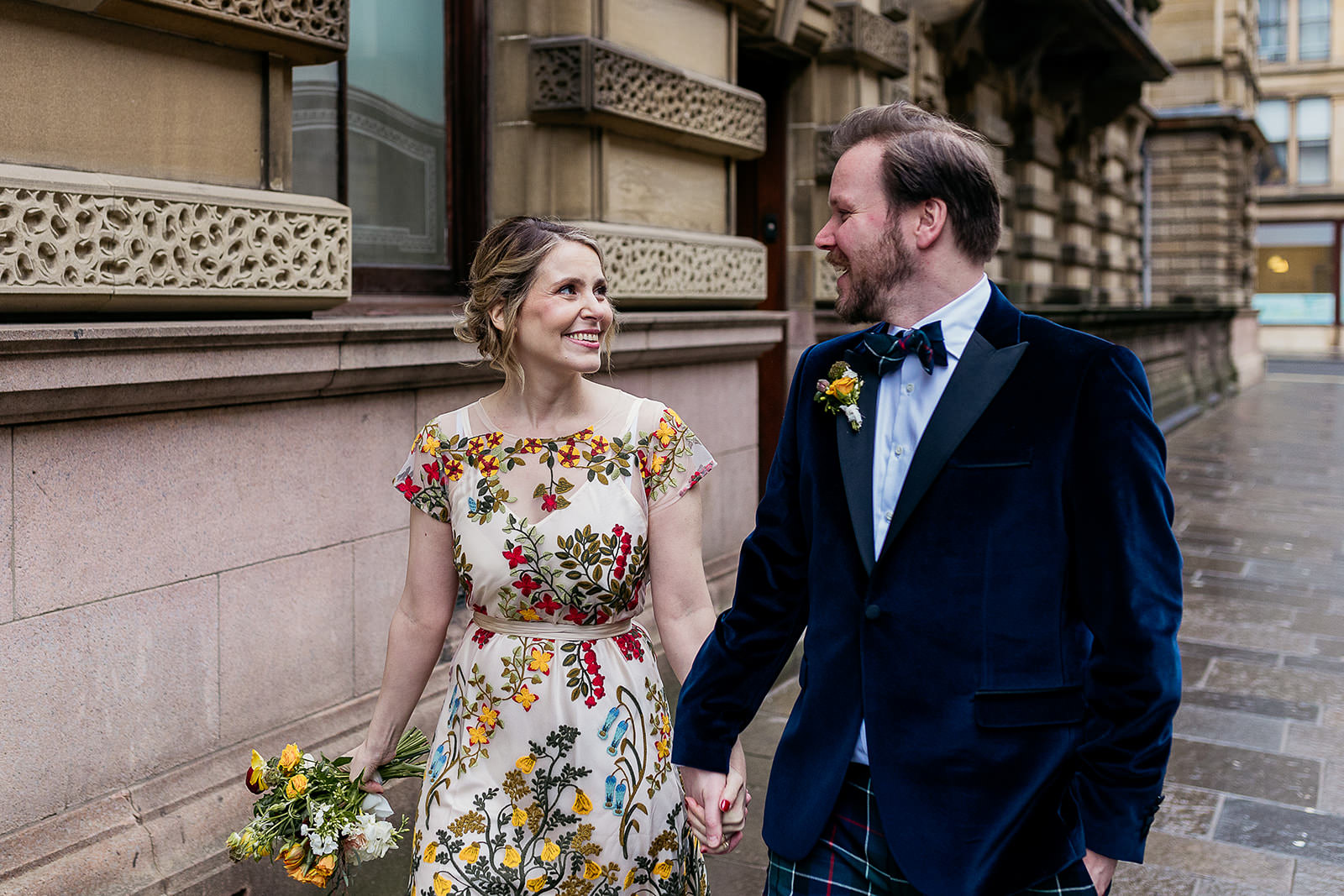 a bride and groom are walking down a street in Glasgow city centre. They are holding hands and looking at each other. the bride's dress is embroidered with colourful flowers and the groom is wearing tartan trews and a blue velvet jacket.