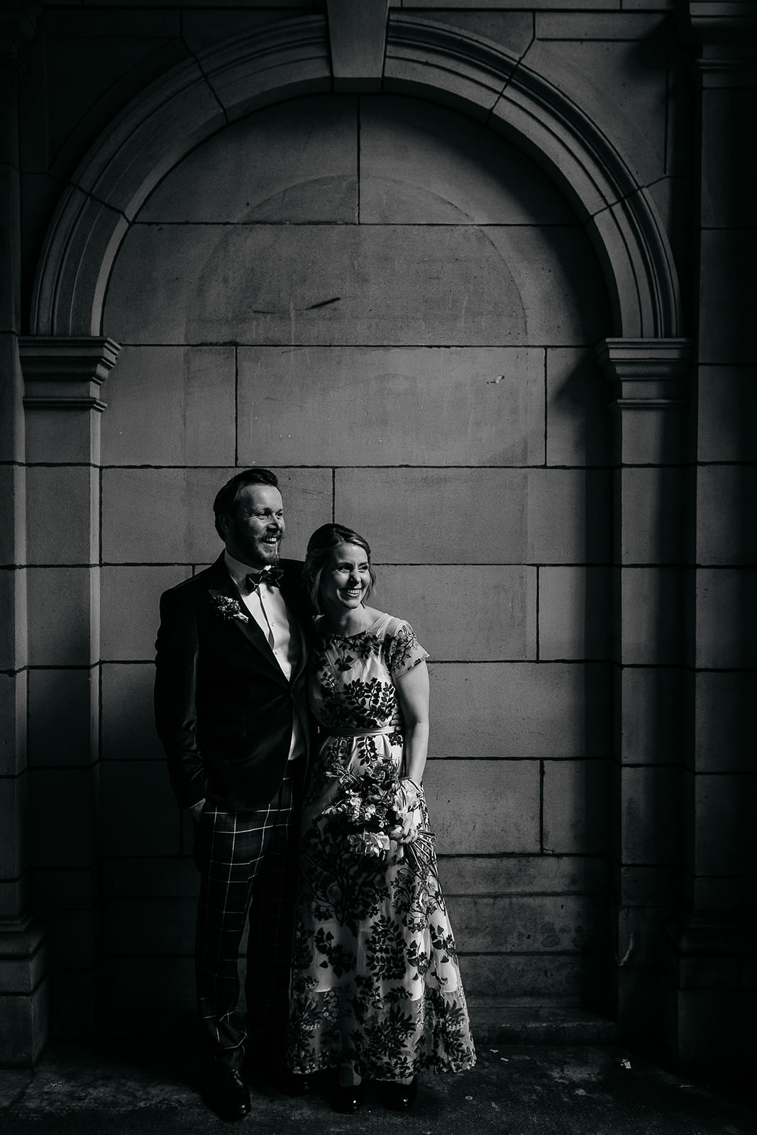 couple are standing together in a secluded spot in front of a stone wall and arch. light is coming in from the right of the frame and highlighting the couple. there is black metal wrought iron gate seen with an ornate design.