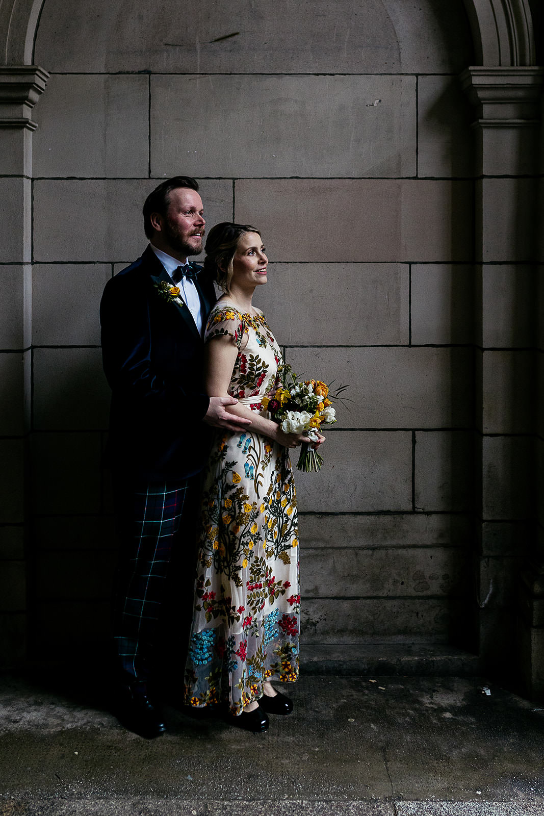 couple are standing together in a secluded spot in front of a stone wall and arch. light is coming in from the right of the frame and highlighting the couple. there is black metal wrought iron gate seen with an ornate design.