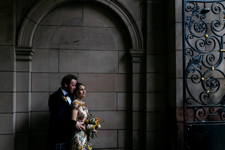 couple are standing together in a secluded spot in front of a stone wall and arch. light is coming in from the right of the frame and highlighting the couple. there is black metal wrought iron gate seen with an ornate design.
