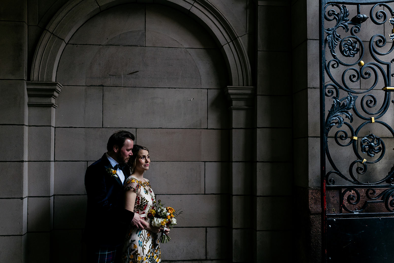 couple are standing together in a secluded spot in front of a stone wall and arch. light is coming in from the right of the frame and highlighting the couple. there is black metal wrought iron gate seen with an ornate design.
