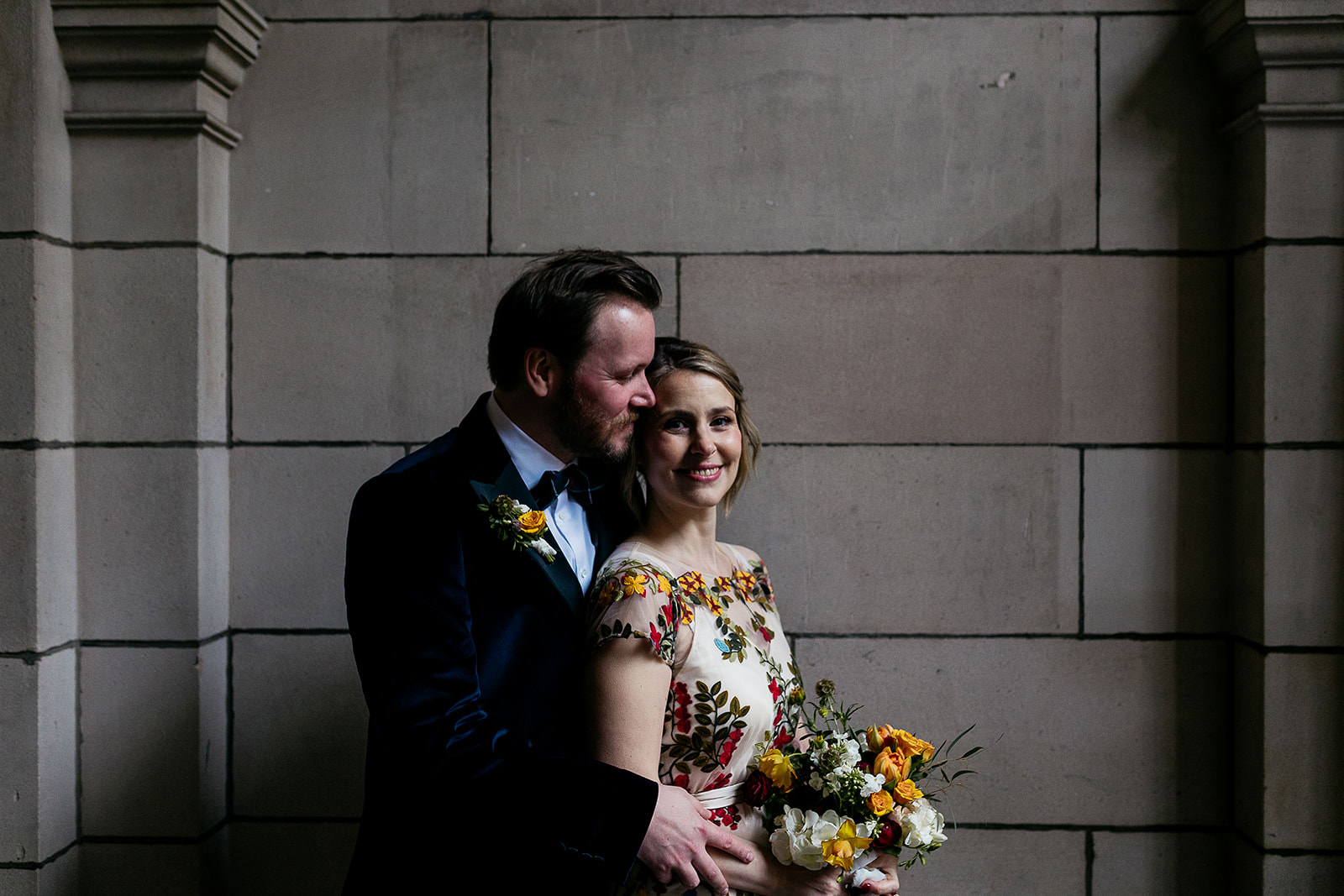 couple are standing together in a secluded spot in front of a stone wall and arch. light is coming in from the right of the frame and highlighting the couple. there is black metal wrought iron gate seen with an ornate design.