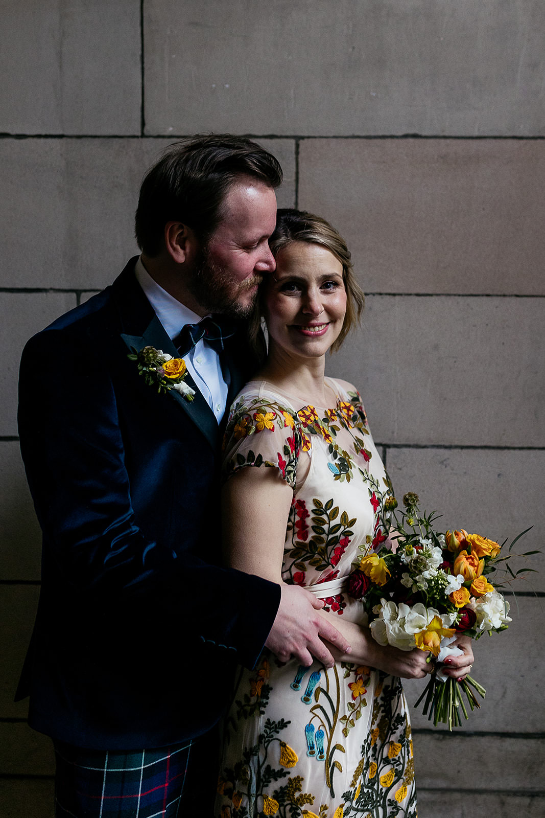couple are standing together in a secluded spot in front of a stone wall and arch. light is coming in from the right of the frame and highlighting the couple. there is black metal wrought iron gate seen with an ornate design.