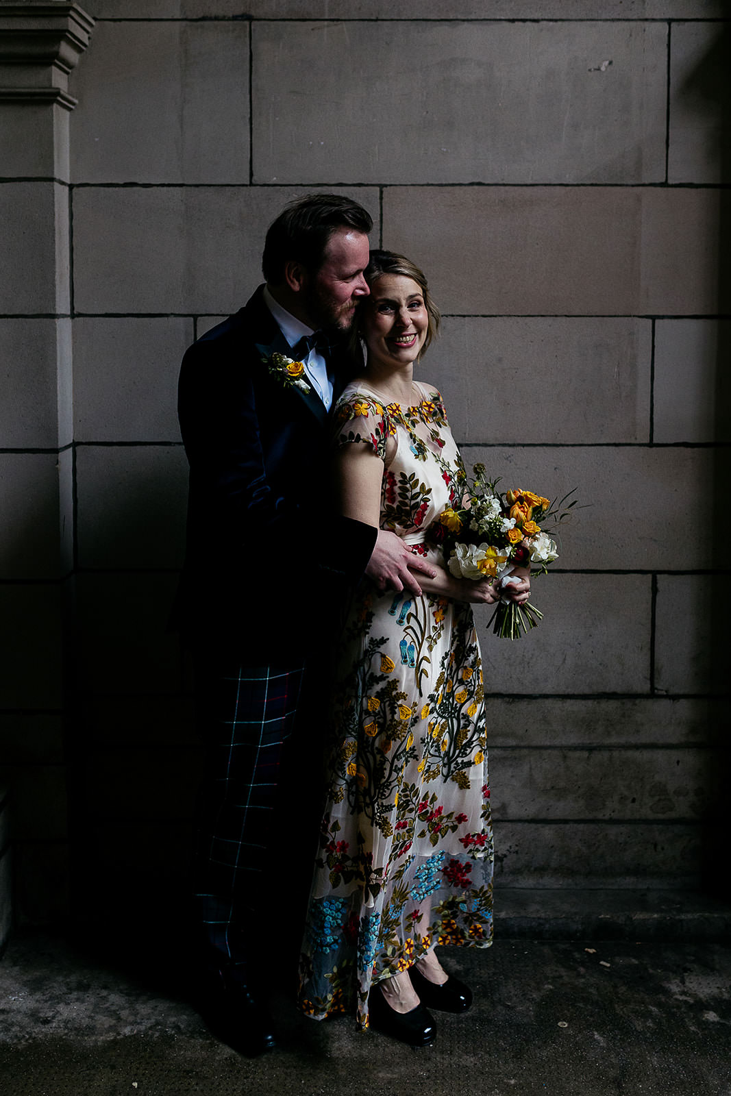 couple are standing together in a secluded spot in front of a stone wall and arch. light is coming in from the right of the frame and highlighting the couple. there is black metal wrought iron gate seen with an ornate design.
