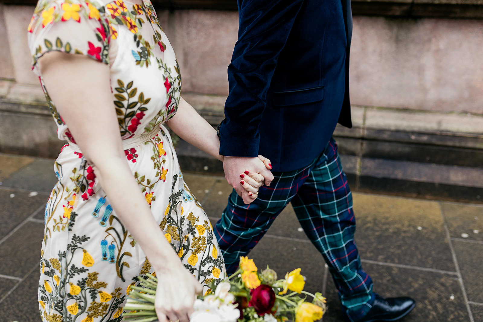a bride and groom are holding hands and walking. the bride nails are painted red and her dress fully embroidered with colourful flowers. the groom is wearing tartan trews and a blue velvet jacket.
