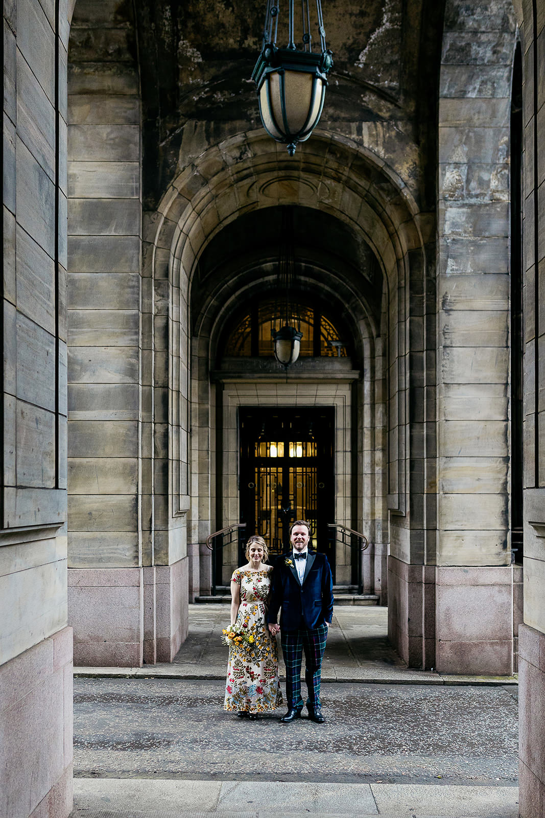 a bride and groom are standing side by side holding hands. they are standing underneath a set of arches and there is light coming from a door behind them.