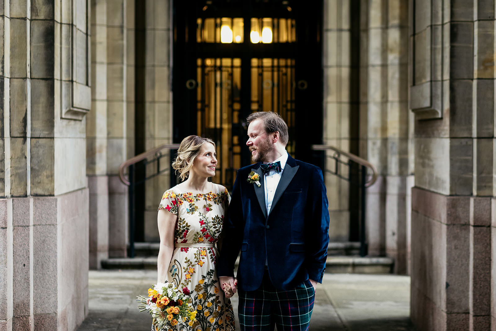 a bride and groom are standing side by side holding hands. they are surrounded by stone arches and there is light coming from a door behind them.