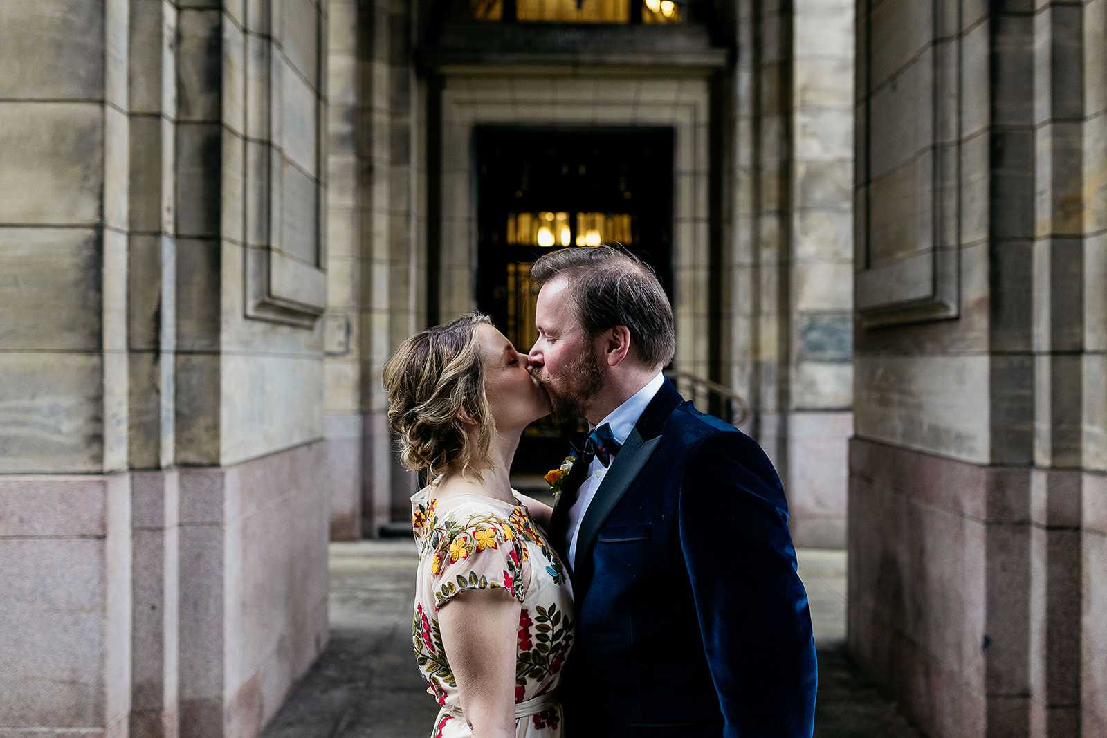 a bride and groom are standing facing each other kissing. they are standing underneath a set of stone arches and there is light coming from a door behind them.