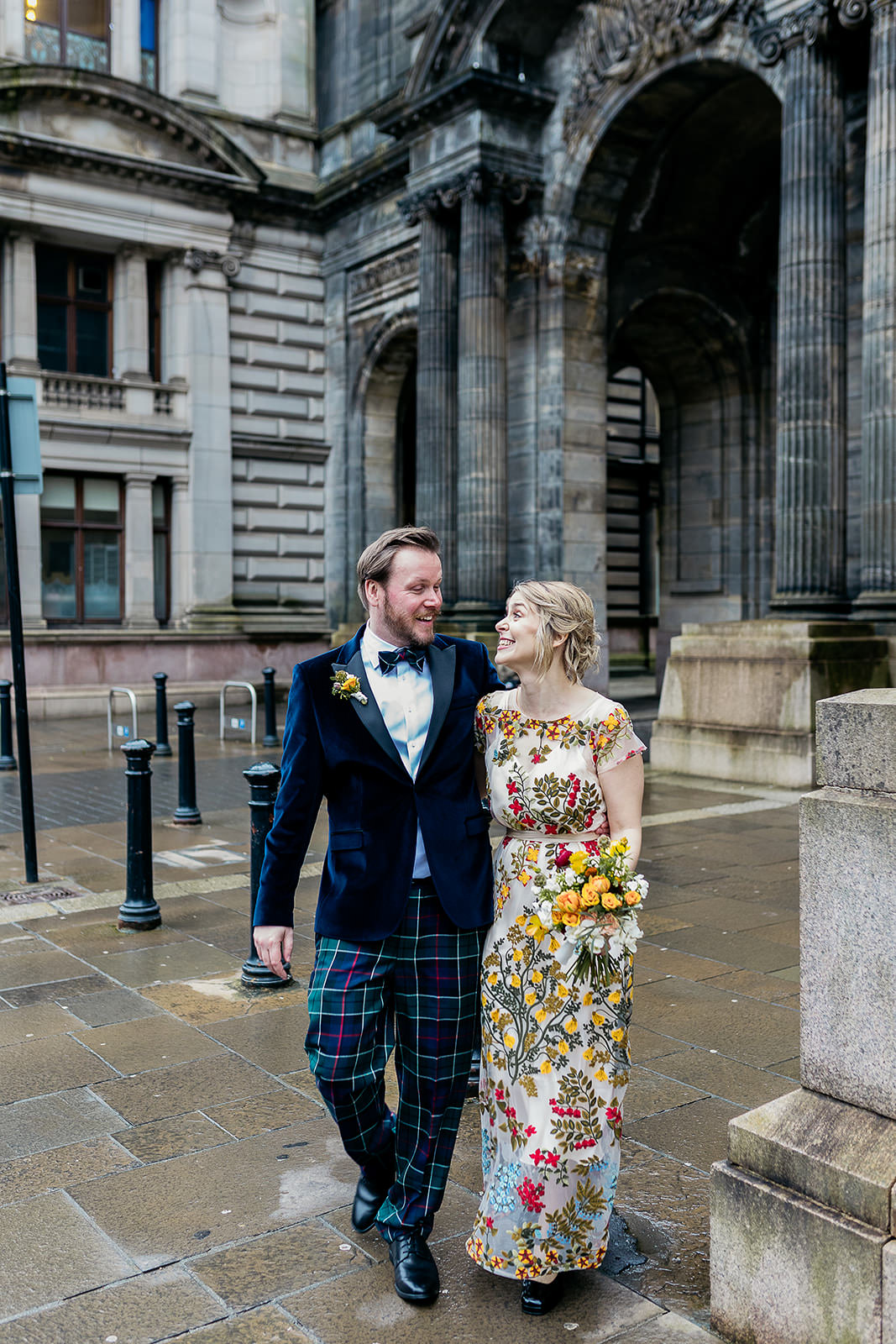 a bride and groom are walking side by side and holding each other close. they have their arms around each other and are walking away from a set of stone arches.